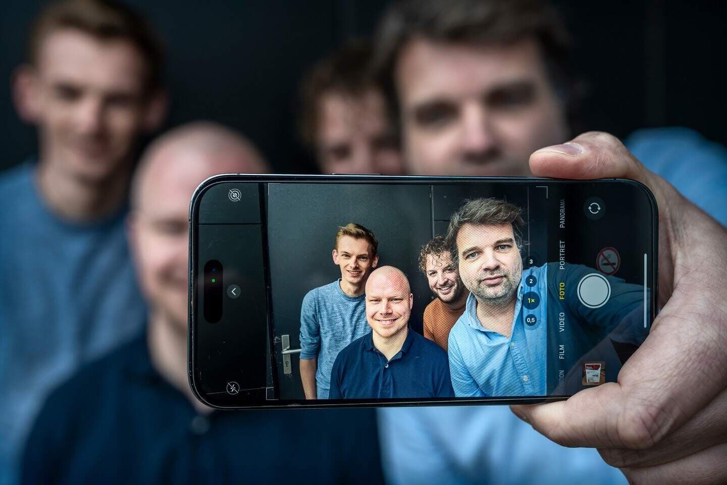 Vier leden van het app-team maken een selfie, gefotografeerd door het scherm van de telefoon. Sebastiaan staat achteraan, met krullen.